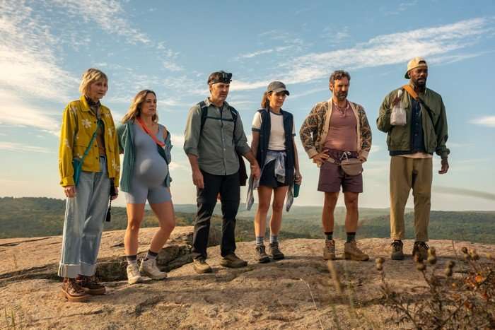 THE FOUR SEASONS, SEASON 2 Six friends stand together on a rocky hilltop during a hiking trip, looking ahead with concerned or uncertain expressions. The group are dressed in casual outdoor clothing, including hiking boots, jackets and backpacks, with one man wearing a head-mounted camera. Rolling hills and a bright blue sky stretch out behind them, creating a scenic backdrop.