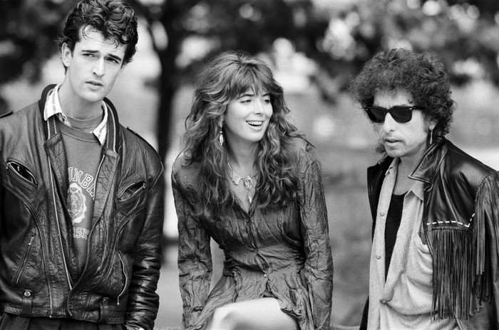 Hearts of Fire Rupert Everett, Fiona Flanagan and Bob Dylan standing in a park wearing casual clothes, Everett in an oversized leather jacket and Dylan wearing sunglasses
