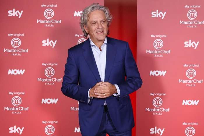 Giorgio Locatelli stood wearing a blue suit and a white shirt, smiling ahead as he poses in front of a red background.