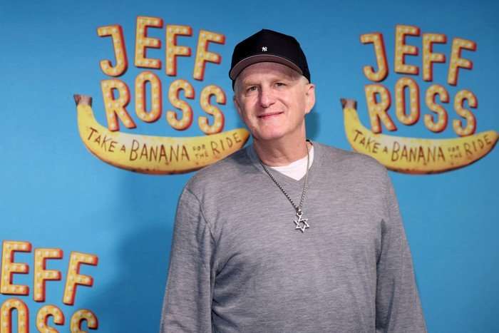 the-traitors-US Michael Rapaport stood wearing a grey jumper and black baseball cap, smiling ahead.