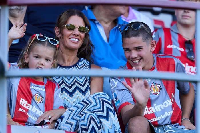 Rebekah Vardy in a zig-zag patterned dress and sunglasses watching a football game with two children dressed in team kit.