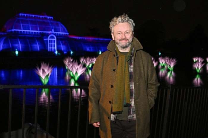 Michael Sheen stood in front of a light display, smiling ahead.