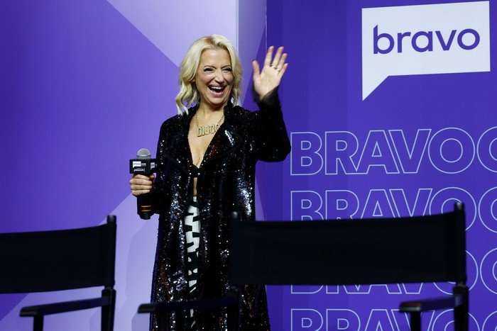 the-traitors-US Dorinda Medley smiling ahead, holding a microphone with one hand and waving with the other.