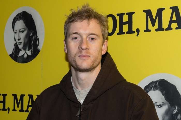 James Acaster stood in front of a yellow background lookinf ahead. He is wearing a black hoodie.