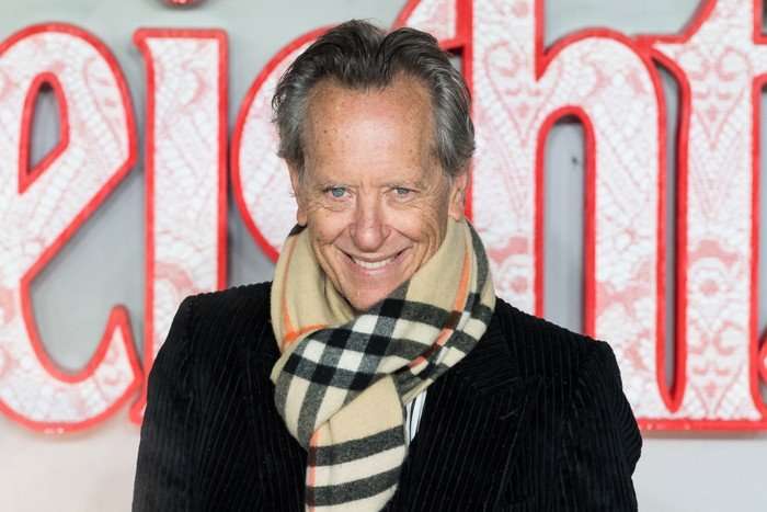 Richard E Grant stood in front of a 'Wuthering Heights' background smiling ahead as he wears s scarf and black trench coat.