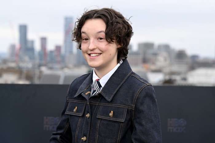 Bella Ramsey smiling ahead on a London rooftop, wearing a black denim jacket and white shirt and tie.