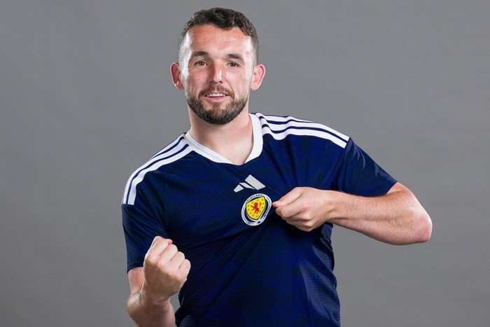Scotland World Cup 2026 Scotland midfielder John McGinn, in blue football kit, pumps his fist and pulls at the crest on his shirt in front of a grey background.