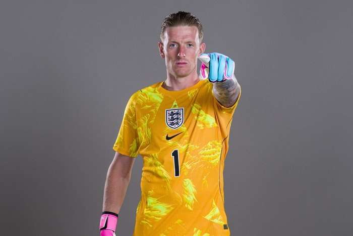 Jordan Pickford England goalkeeper Jordan Pickford, in a yellow kit, points at the camera in front of a grey background.