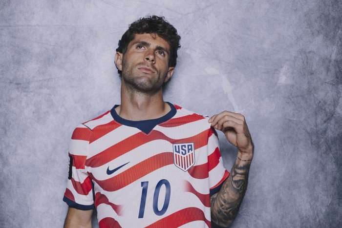 World Cup 2026 USA USA attacker Christian Pulisic, in red and white football kit, pulls on his shirt and looks up as he stands in front of a grey background.