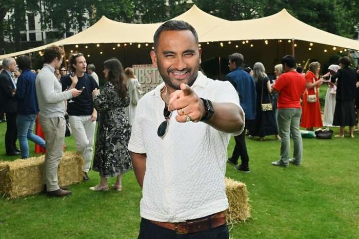 Amol Rajan wearing a white polo shirt and black trousers, smiling as he points his finger ahead of him.