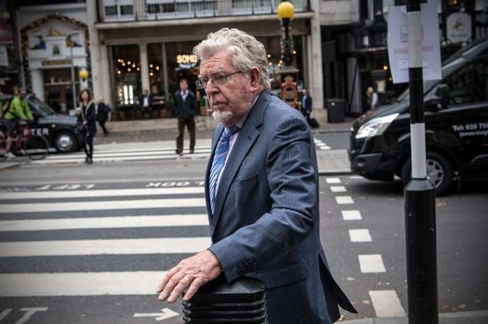 Rolf Harris walking by a zebra crossing, wearing a blue suit.