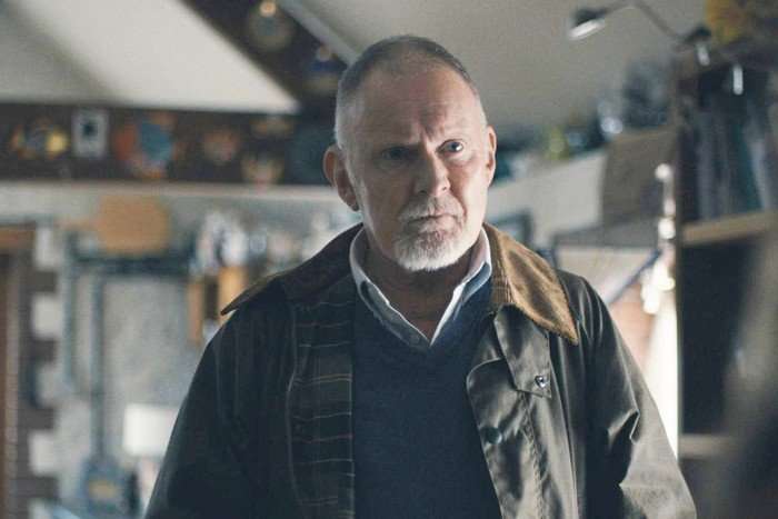 An older man with closely cropped grey hair and a short white beard stands indoors, looking intently toward someone off-camera. He wears a collared shirt layered under a dark sweater and a weathered jacket with a corduroy collar. The room around him is softly lit and slightly cluttered, with shelves, framed items, and household objects blurred in the background, creating a serious and reflective atmosphere.