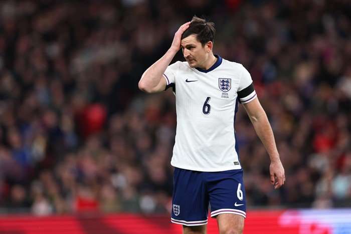 Harry Maguire Harry Maguire, in white and blue England football kit, flattens his hair with his right hand.