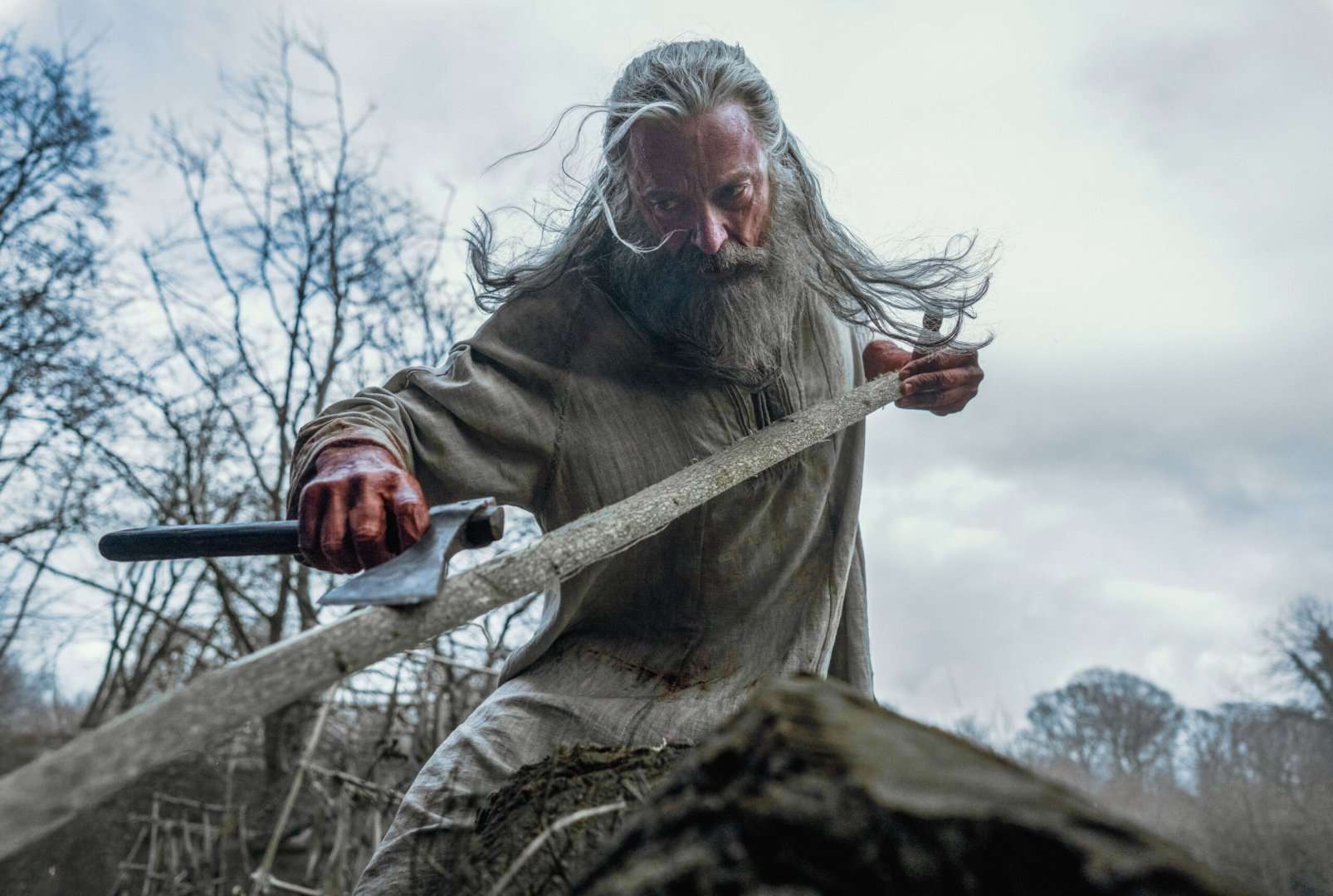 Hugh Jackman sculpting a wooden bow with an ax in the A24 film, The Death of Robin Hood.