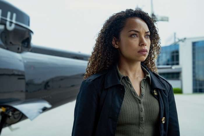 Logan Browning as Agent Sarah Greer in I Will Find You standing outside on an airfield with a helicopter behind her, looking at something seriously.
