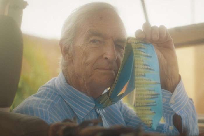 Jon Snow: A Last Big Story Jon Snow in a vehicle holding up a blue and yellow-patterned tie to the camera.
