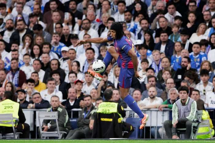 Jules Kounde Jules Kounde, in red and blue Barcelona kit, jumps to take down a football with his foot with Real Madrid fans in the background.