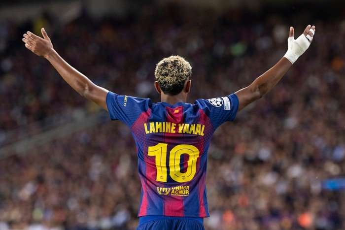 Lamine Yamal, in blue and red striped Barcelona kit, holds both arms up in the air as he faces away from the camera with his name and number on the back of his shirt.