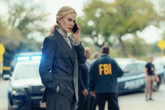 Lioness_302_RG_1016_0539_RT_f Nicole Kidman in Lioness, speaking on the phone outside with a police car and a man wearing an FBI jacket behind her.