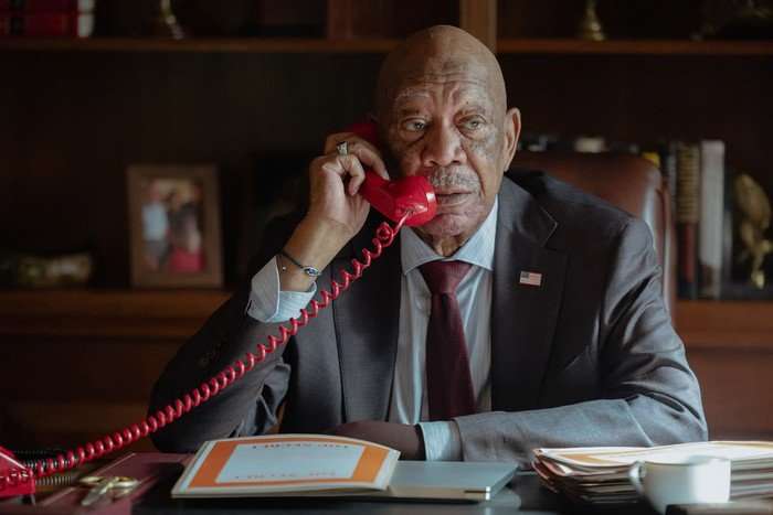 Lioness_304_LS_0112_0210_RT_f Morgan Freeman in Lioness, wearing a suit, sat behind a desk and speaking on a red landline phone.