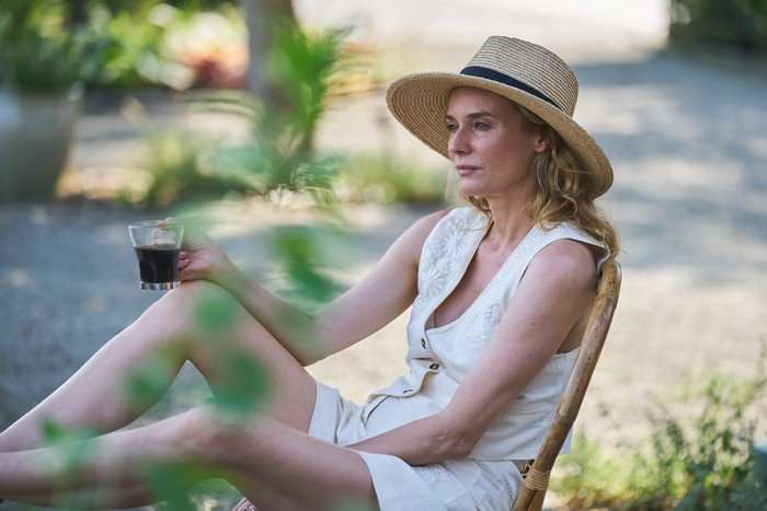Little Disasters SE 1 EP 2 Diane Kruger as Jess Carrisford in Little Disasters wearing a sun hat and white outfit, sitting on a garden chair with her legs up on a ledge and holding a glass of wine.