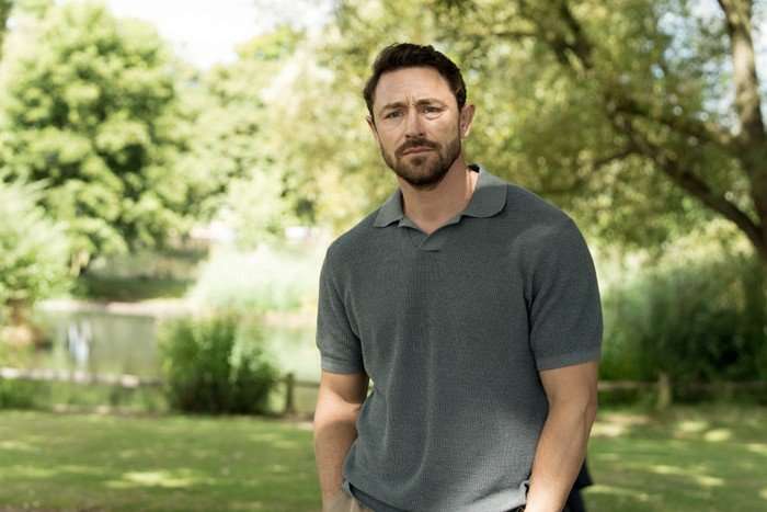 Little Disasters SE 1 EP 3 JJ Feilds as Ed Carrisford in Little Disasters looking sadly across the park, wearing a polo shirt and standing with his hands in his pockets.