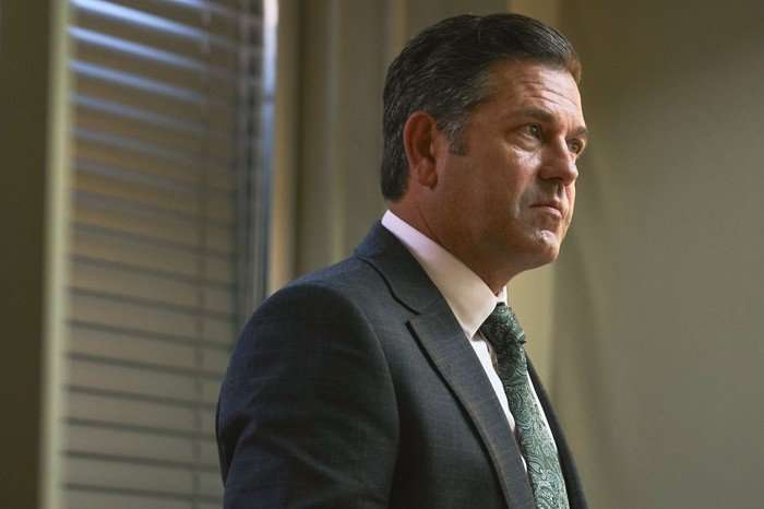 Little Disasters SE 1 EP 5 Patrick Baladi as Andrew Hinman in Little Disasters wearing a suit and standing up in a room.