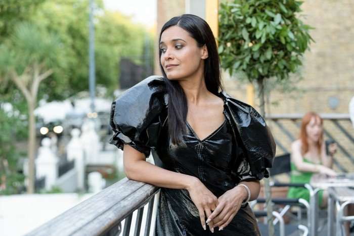 Little Disasters SE 1 EP 3 Shelley Conn as Charlotte Hinman in Little Disasters, wearing an exaggerated black shiny dress and leaning on a balcony, staring mischievously to the side.