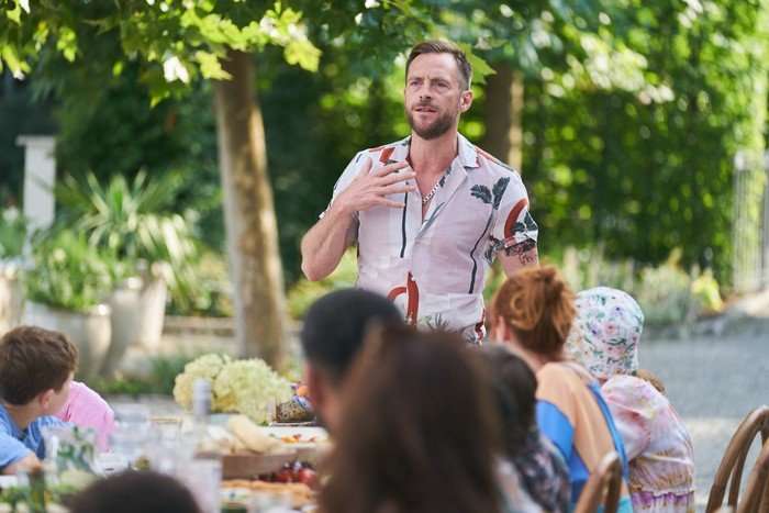 Little Disasters SE 1 EP 2 Stephen Campbell Moore as Rob Baird in Little Disasters standing up in a patterned shirt and giving a speech to a table full of people.