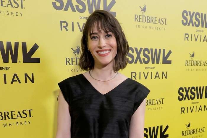Lizzy Caplan smiling in front of a yellow SXSW advertising board.