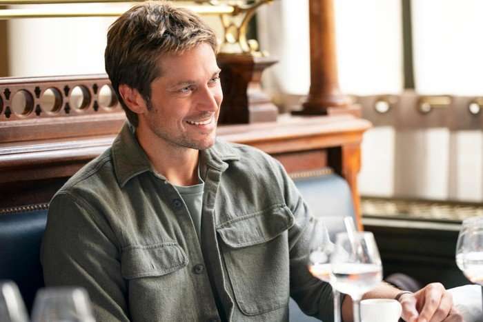 A man with short dark hair smiles while seated at a restaurant table, holding a cup beside a wine glass, with warm wooden interiors and soft daylight in the background.
