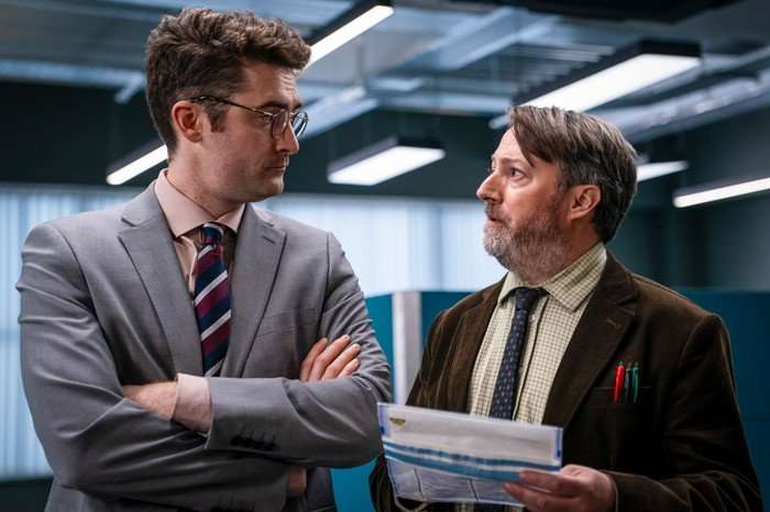 Ben Ashenden as DS Ethan Cole and David Mitchell as John ‘Ludwig’ Taylor in Ludwig. Mitchell is holding a document and they are looking at one another.
