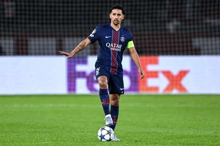 Marquinhos Marquinhos, in blue and red striped PSG football kit, makes a "calm down" gesture as he stands with one foot on a football.