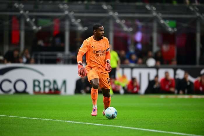 Mike Maignan, in orange AC Milan goalkeeping kit and orange gloves, looks up as he controls a football.