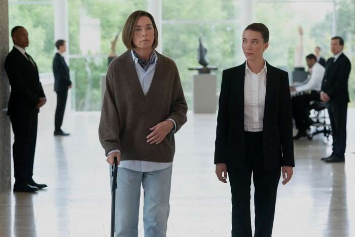 JULIANNE NICHOLSON, NICOLE BRYDON BLOOM Julianne Nicholson walking through an office building lobby wearing a jumper and jeans, walking with a stick while Nicole Brydon Bloom walks next to her in a black suit.