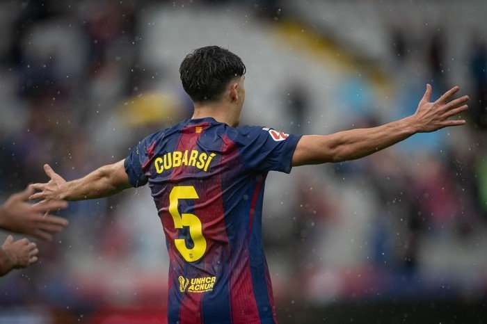 Pau Cubarsi, in red and blue Barcelona kit with his name and number on the back, gestures with both arms wide as he faces away from the camera.