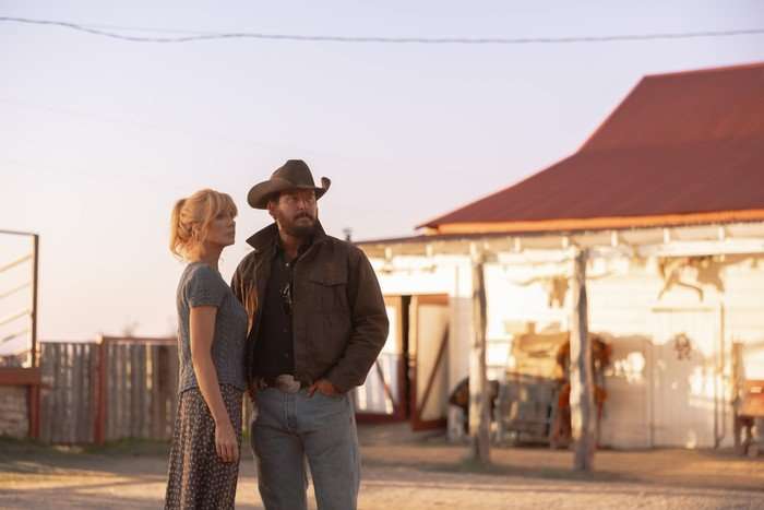 A man and woman stand in front of a white ranch house. Both are wearing jeans and T-shirts, but the man is also wearing a leather jacket and a brown cowboy hat
