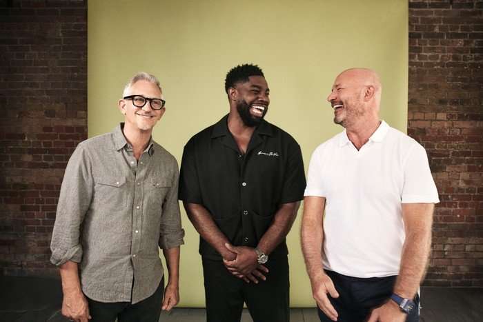 Gary Lineker, Micah Richards, and Alan Shearer smile and laugh in front of a gold background.