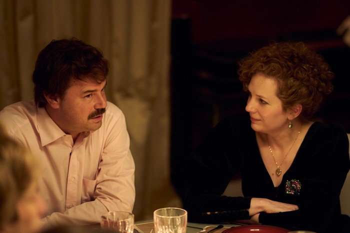 Rivals Danny Dyer as Freddie Jones and Katherine Parkinson as Lizzie Vereker in Rivals. Freddie and Lizzie are sitting in deep conversation at a table and are angled towards each other. Freddie has a moustache and long, cropped hair and is wearing a light pink shirt, while Lizzie has her curly red hair tied up, while wearing a black dress with a v-neckline and a necklace.
