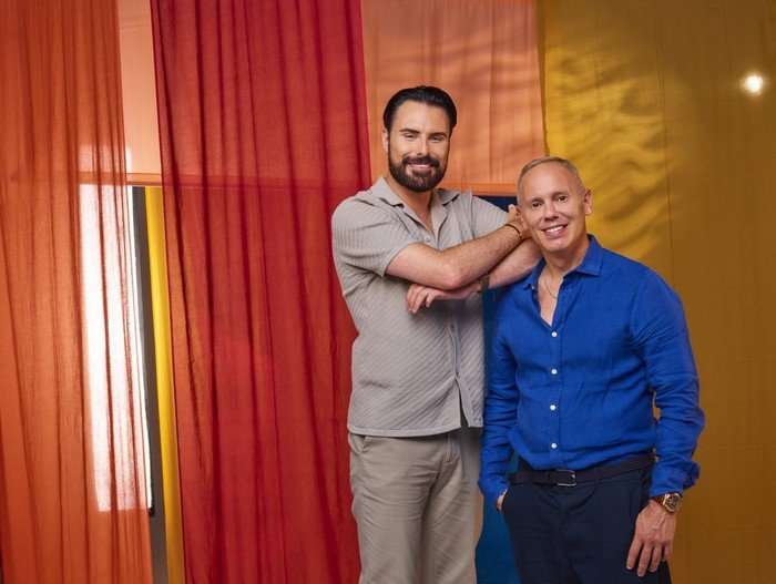 Rylan Clark and Rob Rinder standing in front of a colourful background.