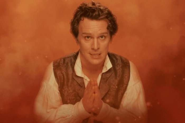 Rogue-Jonathan-Groff-455a225 Jonathan Groff as Rogue with his hands poised in a prayer pose and wearing regency costume while surrounded by orange smoke in the Doctor Who episode Wish World.