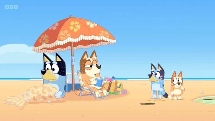 Two animated dogs sit under an umbrella on the beach. One reads a book whilst the other wears a mermaid tail of sand. Two smaller cartoon dogs stand near them.