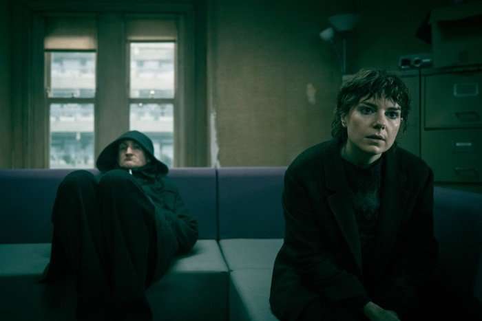 SlowHorses_602_00260F Aimee-Ffion Edwards and Tom Brooke in Slow Horses, sat on a sofa together, both looking ahead. She is leaning forward while he is slouched back with a hood over his head.