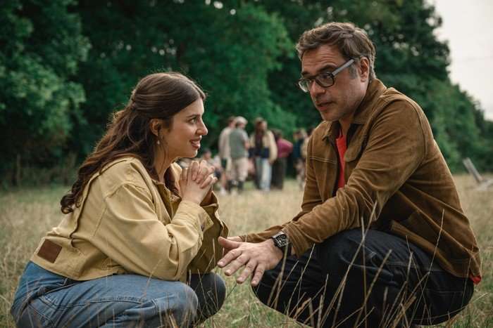 Izzy and Steve share an enthusiastic conversation, while crouching down on the grass in a green park