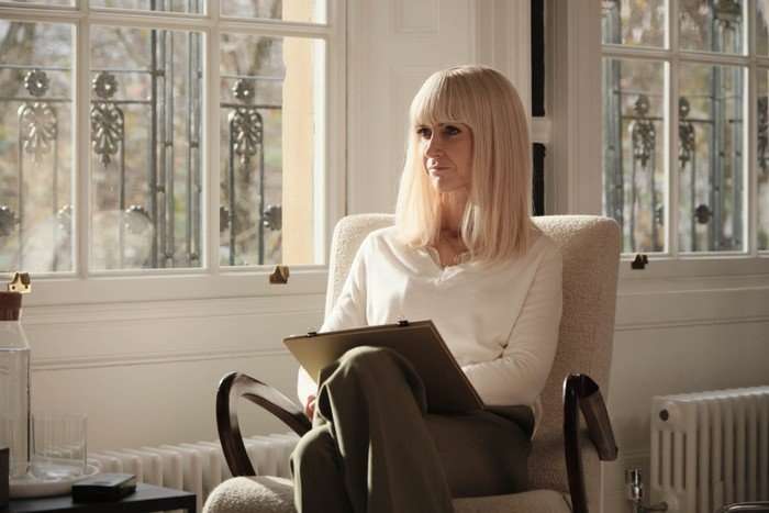 THE CROW GIRL SEASON 2 Katherine Kelly in The Crown Girl season 2, wearing a white jumper, sat in a chair in a room with a clipboard on her lap.