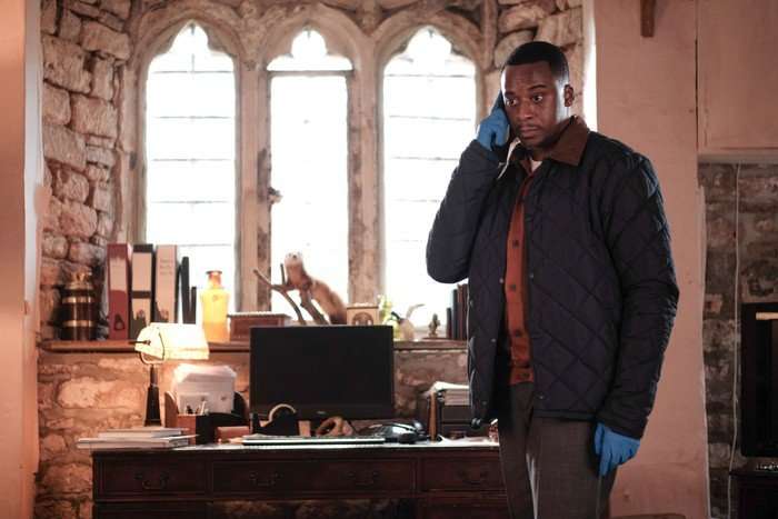 THE CROW GIRL SEASON 2 Elliot Edusah in The Crow Girl, speaking on the phone in a room, wearing blue latex gloves.