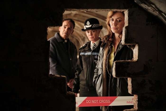 THE CROW GIRL SEASON 2 Eve Myles, Victoria Hamilton and Peter de Jersey in The Crown Girl, looking through a hole in a wall with police tape across it. Hamilton is wearing a police uniform.