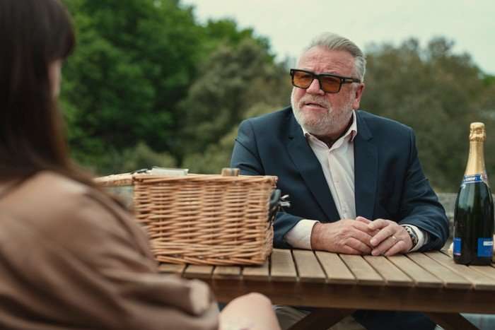 Ray Winstone as Bobby Glass in The Gentlemen. He is sat outside at a table with a basket on it, and is looking at Kaya Scodelario's Suzie Glass, who is off screen