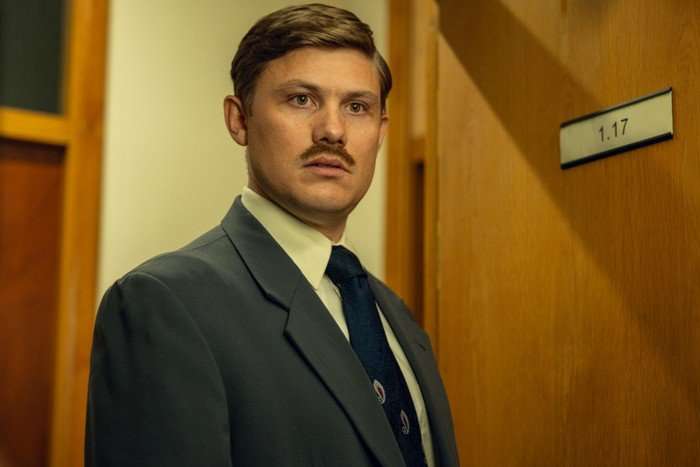 Jonathan Pointing as DC Nick Sparshatt in The Witness standing in a hallway, wearing a suit and shirt, with a door behind him.