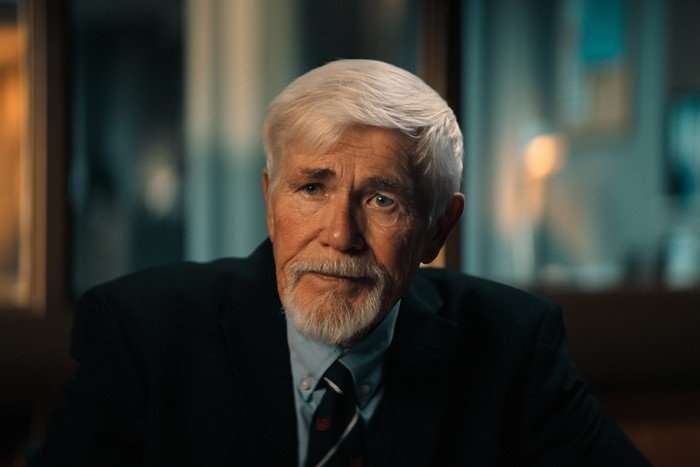 Mickey Banks in The Murder of Rachel Nickell sitting at a desk in a police station, wearing a suit and tie.